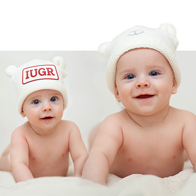 Two babies, one much smaller than the other, with 'IUGR' patch on knit cap