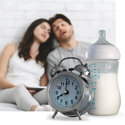 Alarm clock and full baby bottle with exhausted parents in background